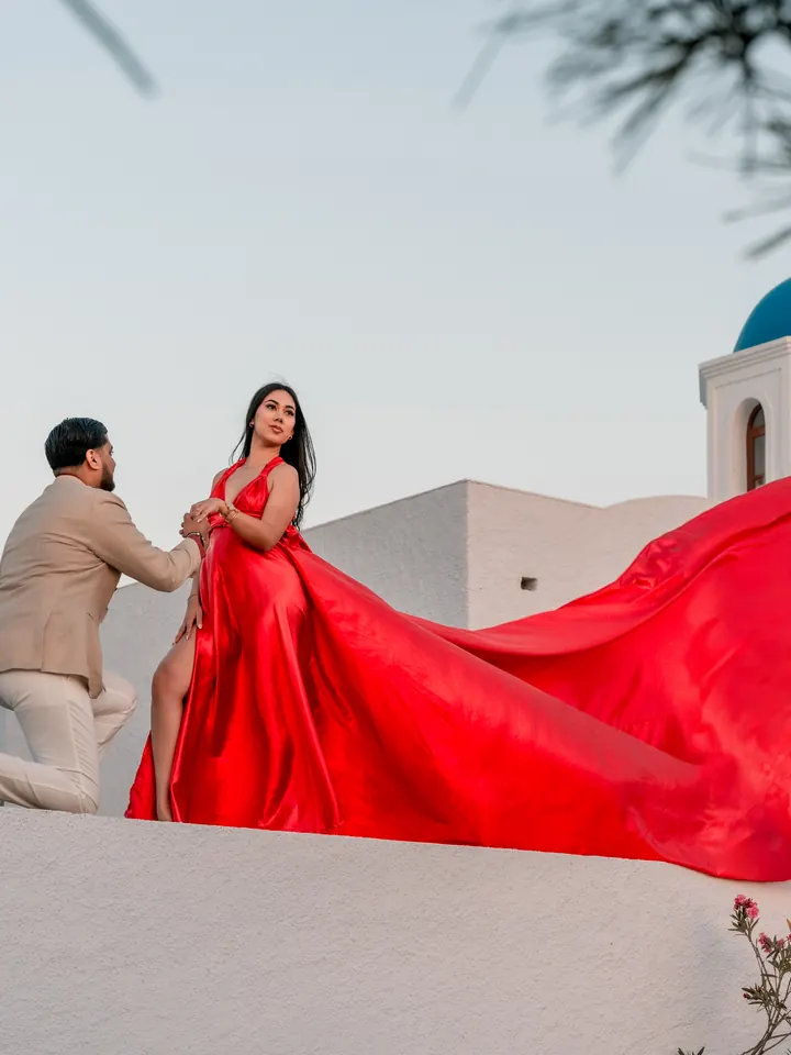A couple pose with a dramatic flowing red dress beside a blue-domed Santorini church